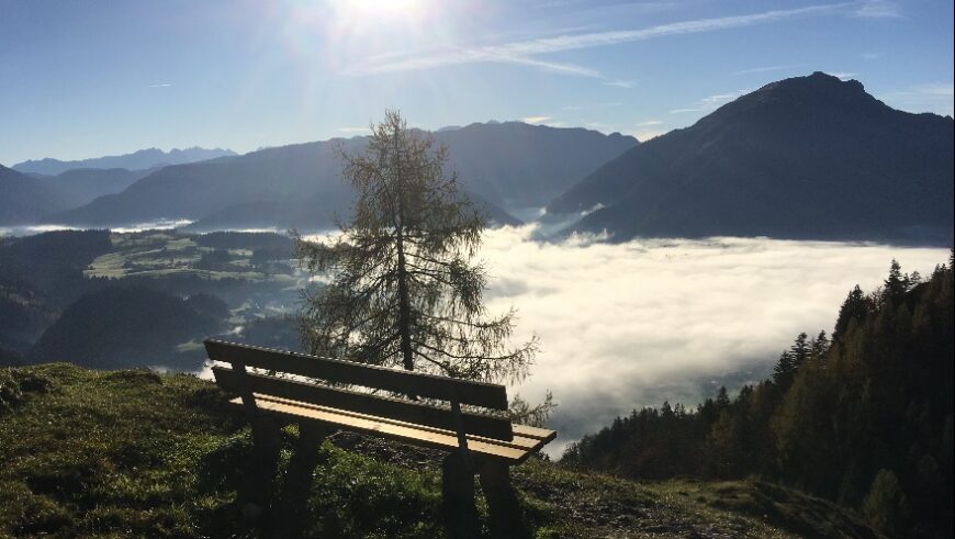 Eine Holzbank steht auf einem grasbewachsenen Hügel. Im Vordergrund befindet sich ein Baum. Die Bank blickt auf ein weites Tal, das von einer dichten Nebeldecke bedeckt ist. Im Hintergrund erheben sich Berge unter einem klaren, sonnigen Himmel. Die Sonne scheint hell am oberen Bildrand.