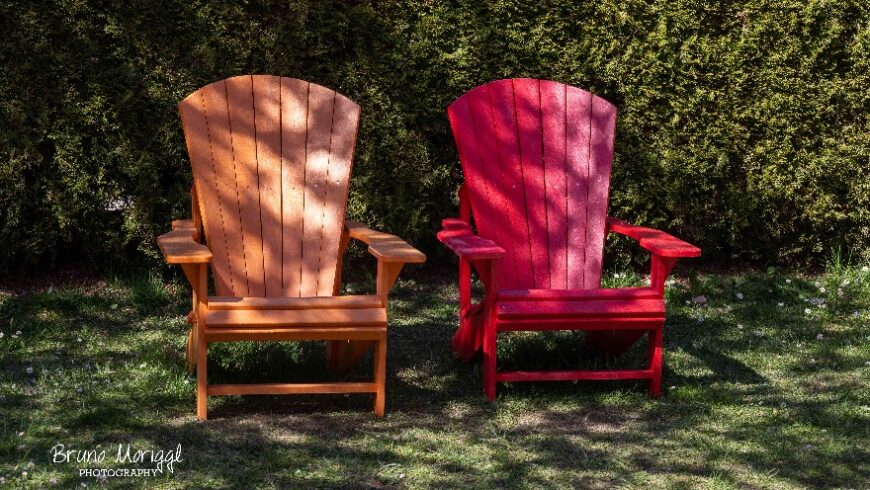 Zwei Adirondack-Stühle stehen im Freien auf einem Rasen. Der linke Stuhl ist in einem hellen Holzton, der rechte ist rot lackiert. Beide Stühle sind von der Sonne beschienen. Im Hintergrund befindet sich eine dichte, grüne Hecke.