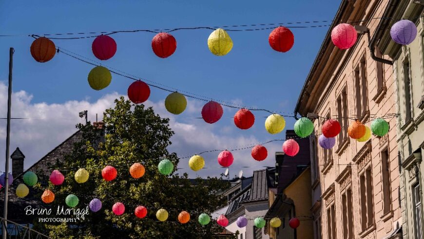 Ein Bild von bunten Lampions, die über eine Straße zwischen Häusern gespannt sind. Die Lampions haben verschiedene Farben, darunter Rot, Pink, Gelb, Grün, Orange, Lila und Weiß. Am oberen Rand sind zwei Leinen mit Lampions zu sehen, die auf eine dritte Leine im unteren Bereich zuführen, die in der Ferne verschwindet. Die Häuser haben helle Fassaden mit Fenstern und Dächern. Ein großer grüner Baum ist auf der linken Seite des Bildes zu sehen. Der Himmel ist blau mit weißen Wolken.
