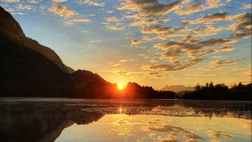 Ein Panoramabild des Sonnenuntergangs über einem See. Die Sonne ist ein leuchtender goldener Punkt, der knapp über dem Horizont steht und einen Sterneneffekt erzeugt. Das Licht der Sonne reflektiert auf der Wasseroberfläche, die von leichten Wellen und Wasserpflanzen bedeckt ist. Auf der linken Seite erheben sich dunkle Berge, deren Silhouette sich scharf vom hellen Himmel abzeichnet.