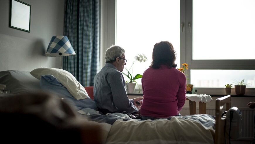 Ein Mann und eine Frau sitzen auf der Bettkante eines Krankenhausbettes und blicken nach draußen in ein helles, großes Fenster. Der Mann mit kurzem, grauem Haar sitzt links, die Frau mit schulterlangem, braunem Haar sitzt rechts. Sie sind beide mit dem Rücken zur Kamera zugewandt.