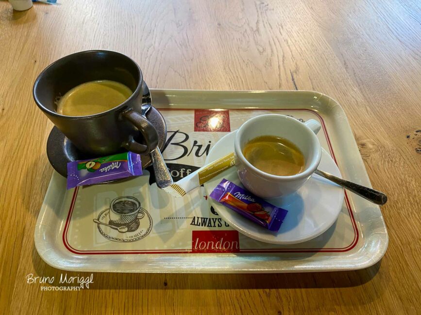 Ein Hochwinkelbild, das zwei Tassen Kaffee auf einem Serviertablett zeigt, das auf einem Holztisch steht. Die eine Tasse ist dunkel und steht auf einer Untertasse. Die andere Tasse ist weiß und kleiner und steht ebenfalls auf einer Untertasse. Jede Tasse hat einen Löffel und ein kleines Stück Schokolade in lila Verpackung. Ein Zuckerpaket liegt zwischen den beiden Tassen.