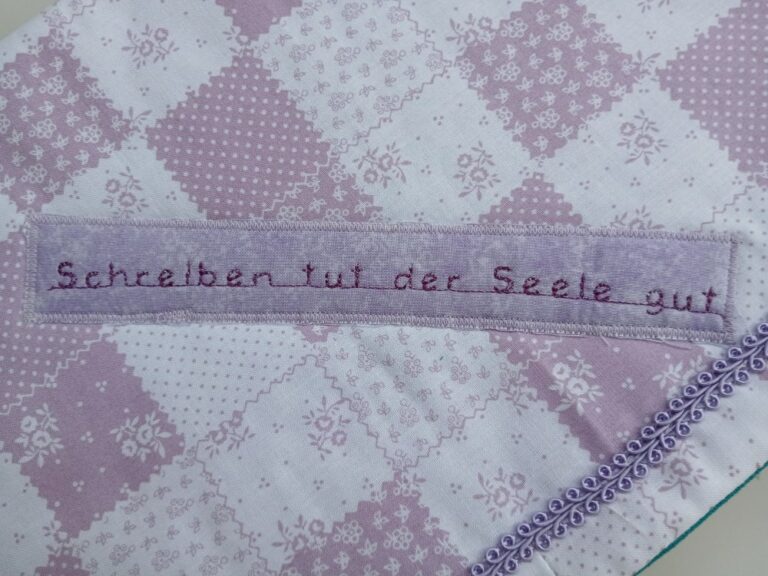 Ein handgenähtes Stoffstück mit einem violett-weißen Patchworkmuster. Darauf ist ein hellvioletter Stoffstreifen aufgenäht, auf dem in lila Schrift die Worte „Schreiben tut der Seele gut“ gestickt sind. Unten rechts verläuft eine dekorative Borte mit kleinen Schleifen.