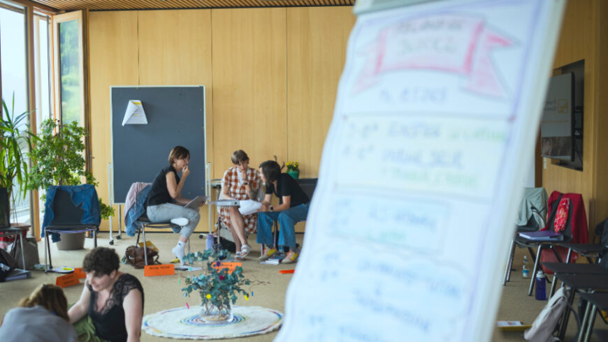 Ein Gruppenfoto, das eine informelle Diskussionsrunde in einem Raum mit Holzwänden zeigt. Drei Personen sitzen auf Stühlen in der Mitte des Bildes und blicken aufeinander. Zwei weitere Personen sind im Vordergrund teilweise sichtbar.